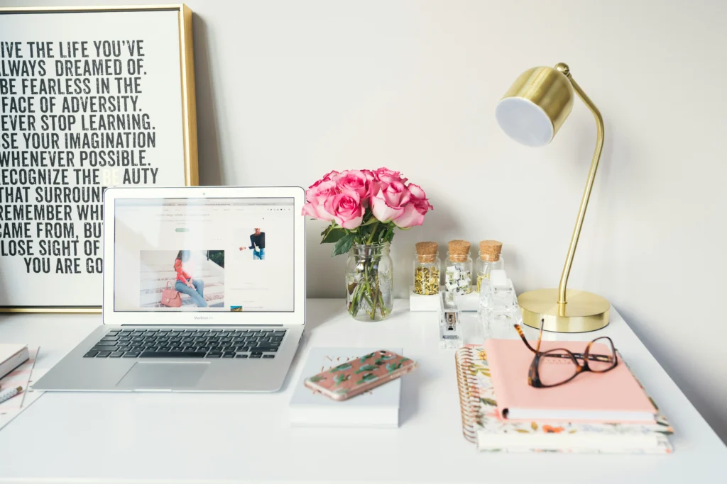 a table with a laptop, desk lamp, a bouquet of pink roses and other things